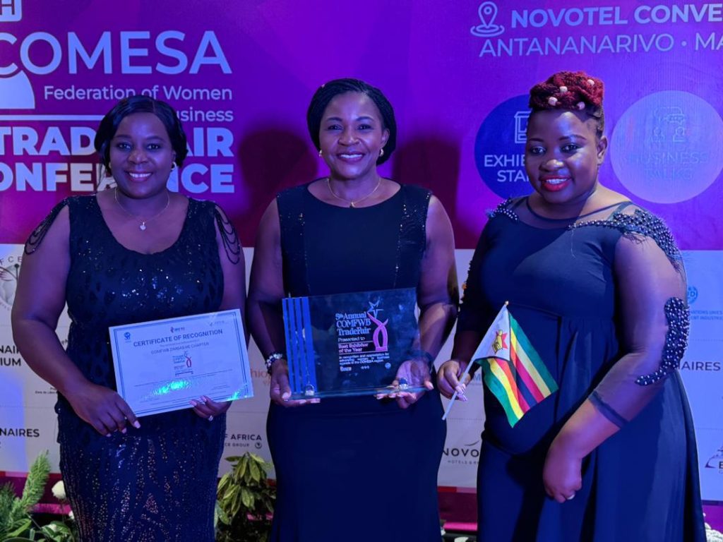 Three women wearing glamorous black dresses standing in front of a COMESA photo backdrop. One holds a certificate of recognition, another the award and the third a small Zimbabwean flag.