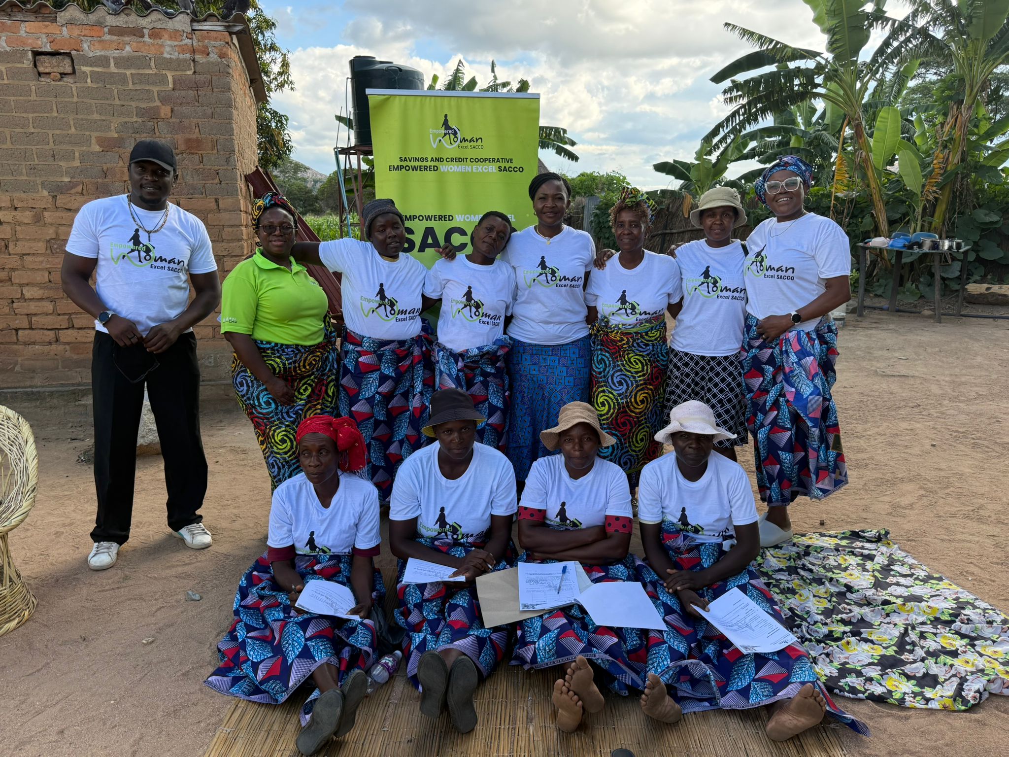 Various women and a man wearing SACCO branded tshirts in a rural area with a SACCO roll up banner behind.